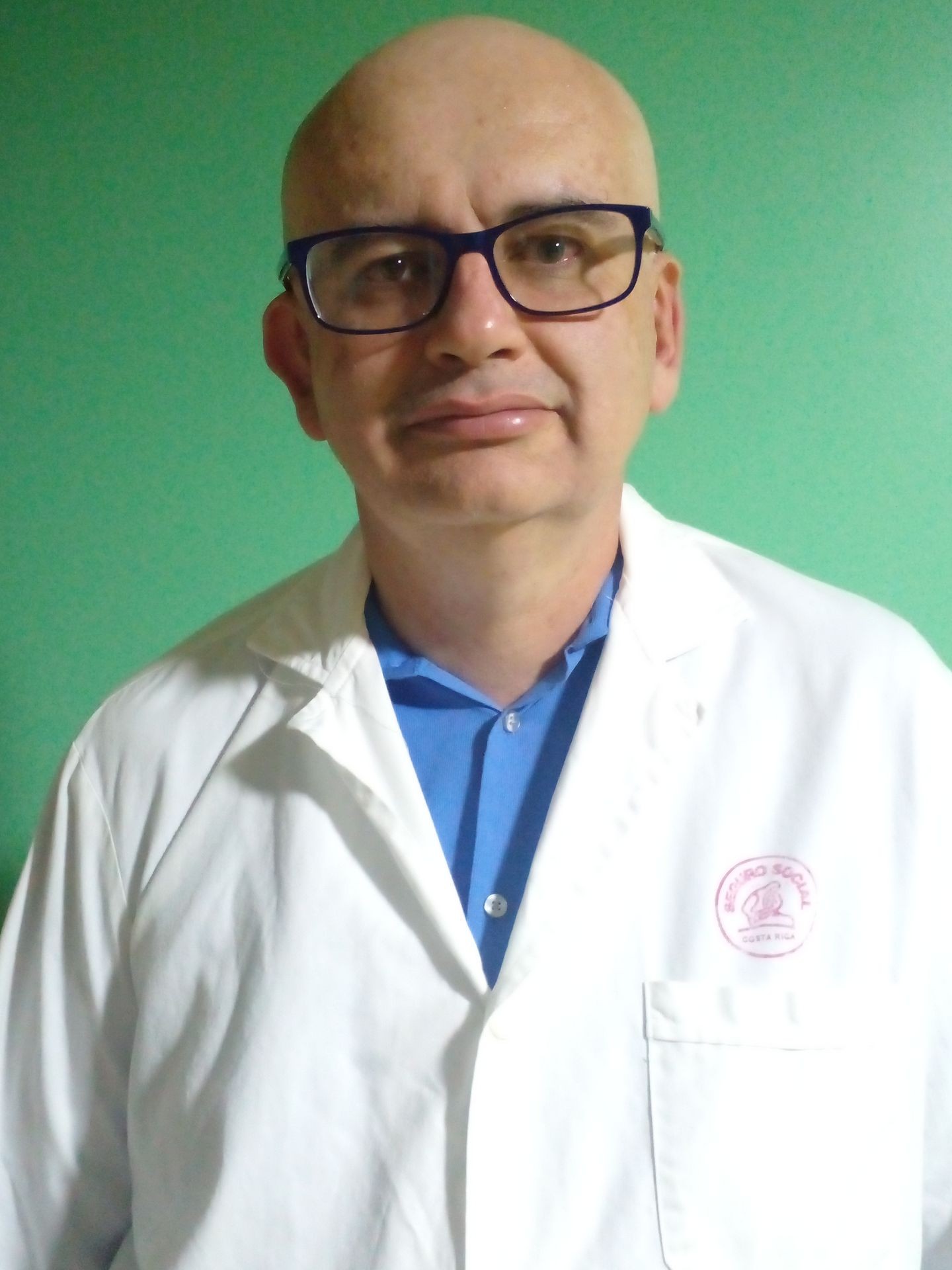 Person wearing a white lab coat over a blue shirt, standing against a green background.