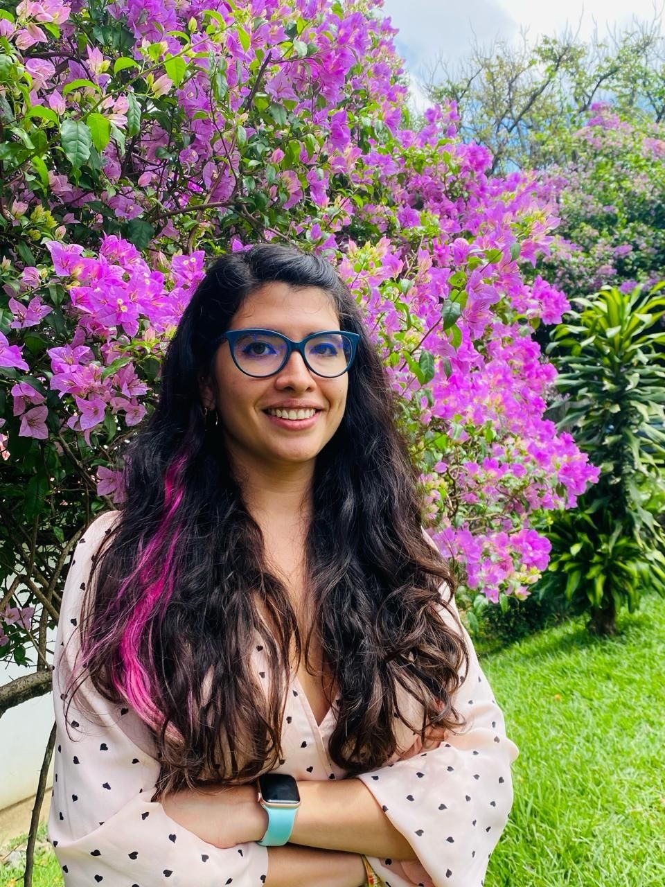 Smiling woman with long hair and glasses standing in front of vibrant purple flowers in a garden.