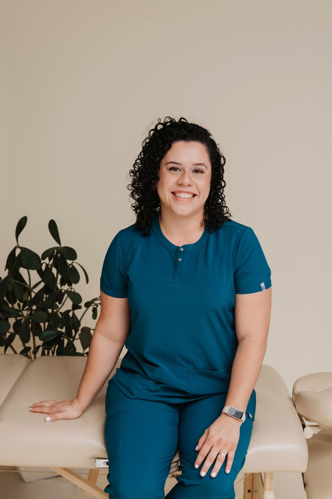 Smiling woman in teal scrubs sitting on a massage table with a plant in the background.